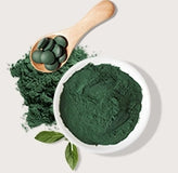 Spirulina powder in a bowl with spirulina tablets on a wooden spoon, surrounded by fresh spirulina leaves.